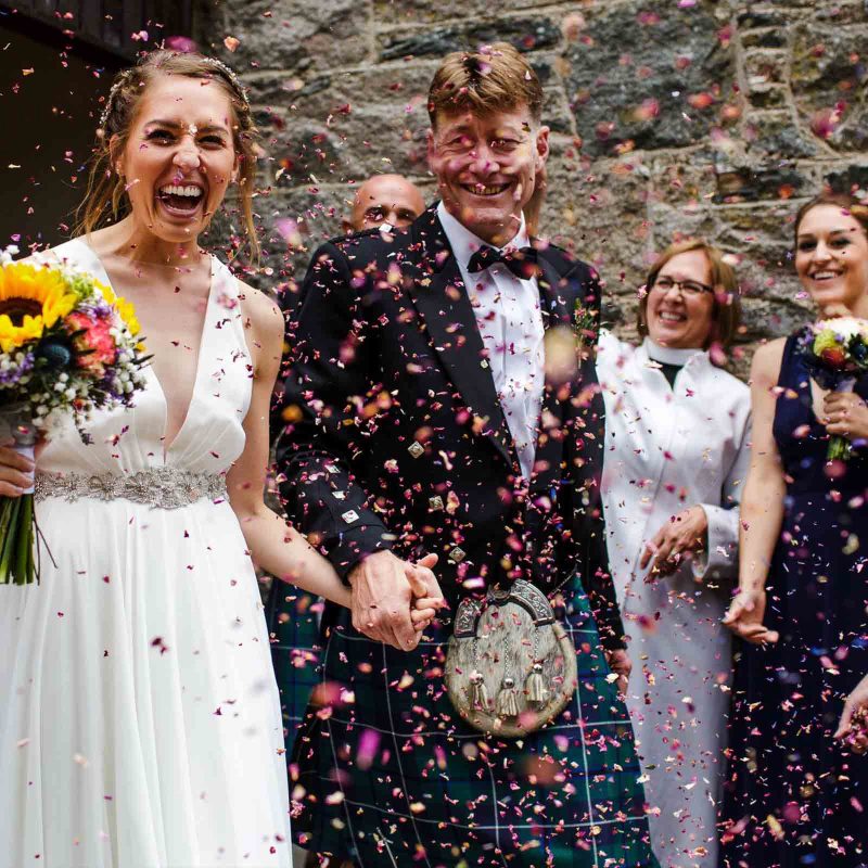 A laughing bride and groom leaving the Kirk in Glen Clova to confetti being thrown over them on their wedding day. Photography by Barry Robb Photography. https://barryrobbphotography.com/