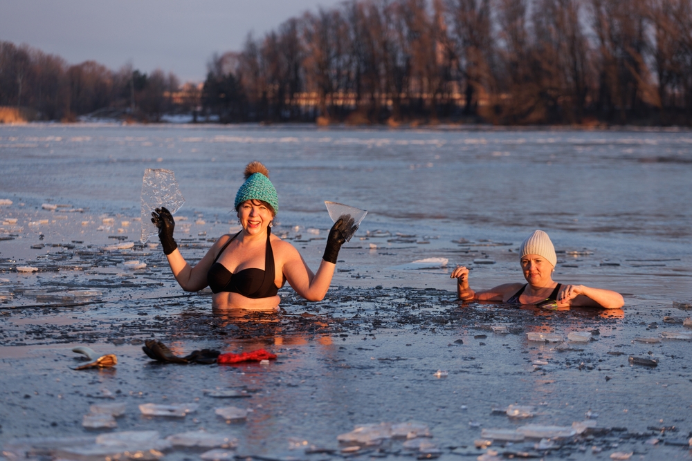 Winter swimming. Two mature woman in frozen lake ice hole. Swimmers wellness in ice. Endorphin booster swim in cold water. Beautiful female body tempering. Cold winter morning landscape.