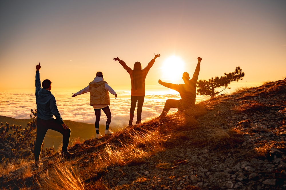 Group of happy friends are standing at mountain top and greeting sunrise or sunset above clouds.