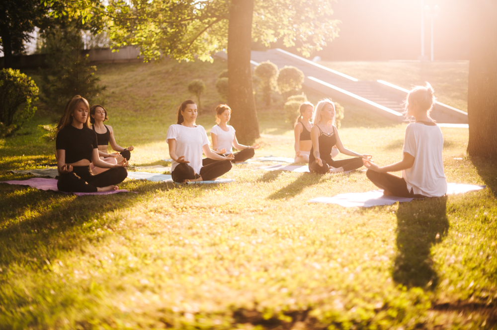 Group of young women practice yoga in park on summer sunny morning under guidance of instructor. Group of calm people is sitting in lotus pose on grass with closed eyes.